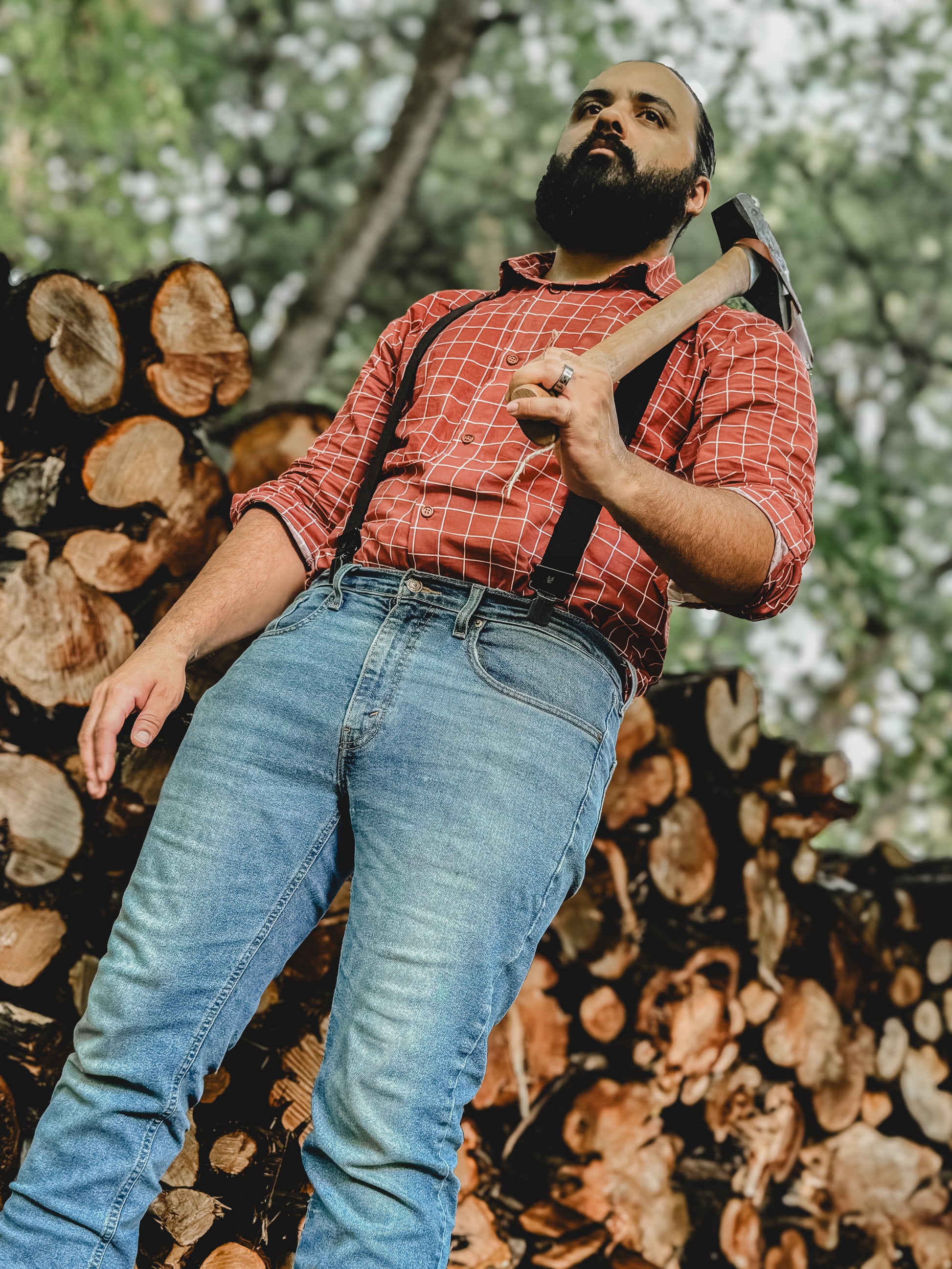 Man in red plaid shirt and blue jeans holding an axe against a background of stacked logs.