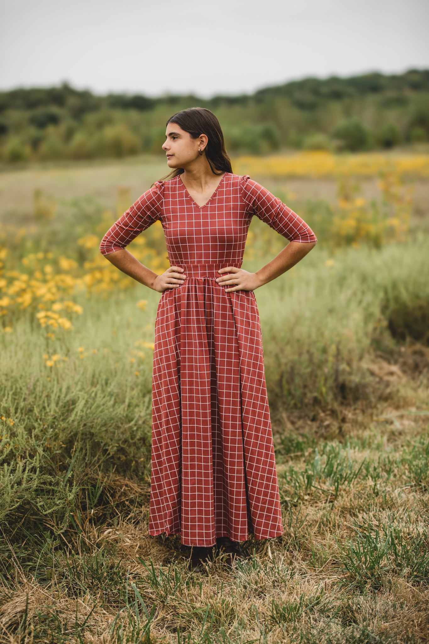 Woman wearing modest copper windowpane plaid maxi dress with 3/4 sleeves and v-neckline, standing gracefully in wildflower field with yellow blooms and lush greenery, showcasing the nursing-friendly design and comfortable cotton spandex fabric ideal for outdoor occasions