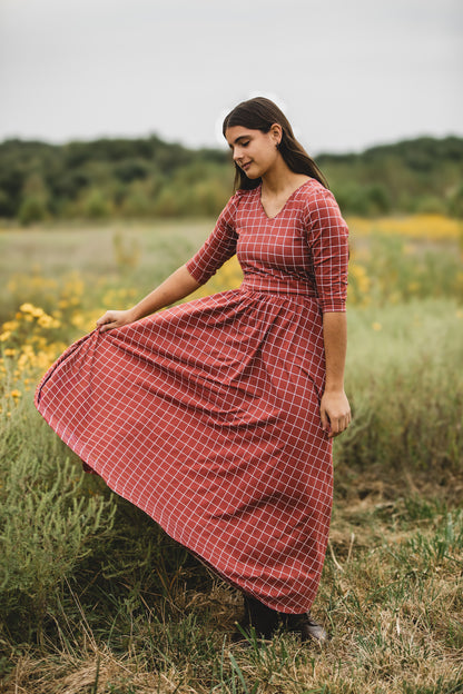 Woman wearing modest copper windowpane plaid maxi dress with 3/4 sleeves and v-neckline, standing gracefully in wildflower field with yellow blooms and lush greenery, showcasing the nursing-friendly design and comfortable fit ideal for outdoor occasions