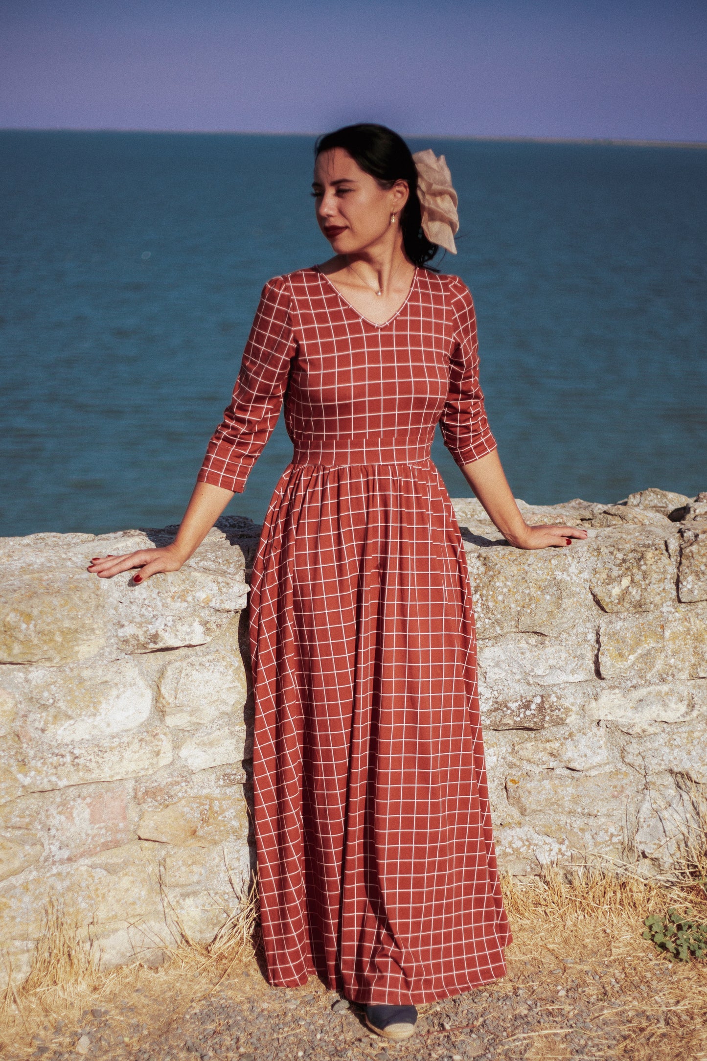 Woman wearing modest copper windowpane plaid maxi dress standing elegantly by rustic stone wall with ocean in background, showcasing the nursing-friendly design with 3/4 sleeves and v-neckline, cotton blend fabric flowing beautifully in coastal breeze perfect for seaside photography and outdoor occasions