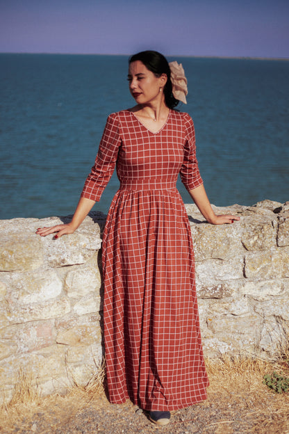 Woman wearing modest copper windowpane plaid maxi dress standing elegantly by rustic stone wall with ocean in background, showcasing the nursing-friendly design with 3/4 sleeves and v-neckline, cotton blend fabric flowing beautifully in coastal breeze perfect for seaside photography and outdoor occasions