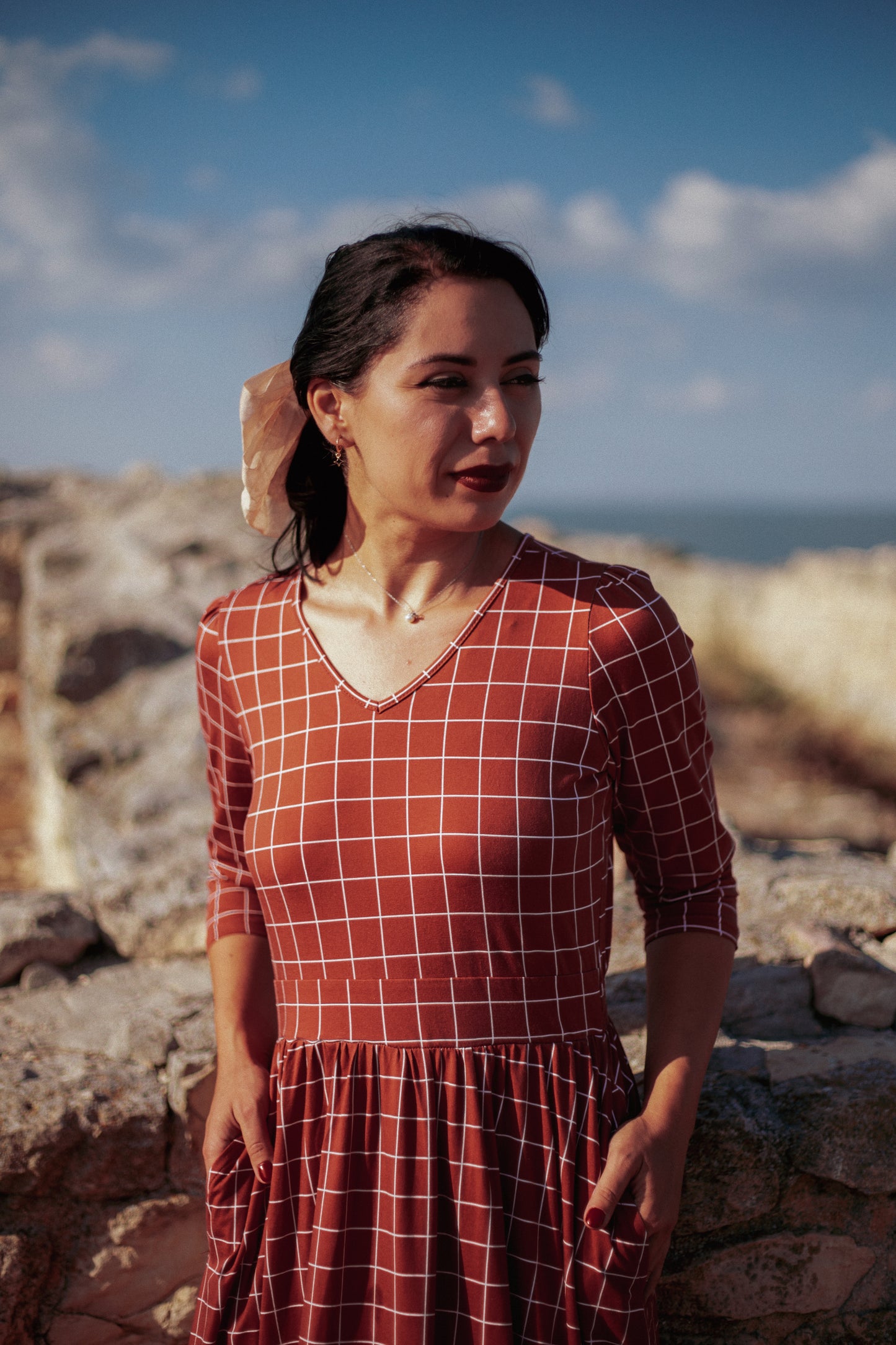 Woman wearing modest copper windowpane plaid maxi dress with 3/4 sleeves and v-neckline, standing gracefully on coastal cliffs with ocean waves and blue sky in background, demonstrating the nursing-friendly design and comfortable cotton spandex fabric perfect for seaside adventures