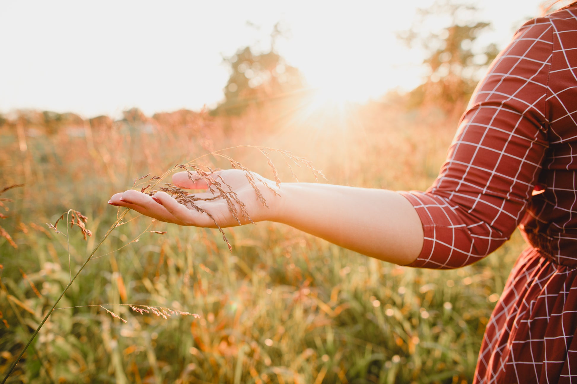 Woman wearing modest copper windowpane plaid maxi dress standing gracefully in sunlit field holding tall grass, showcasing the nursing-friendly design with 3/4 sleeves and v-neckline, cotton blend fabric flowing beautifully in golden hour lighting perfect for maternity and outdoor photography