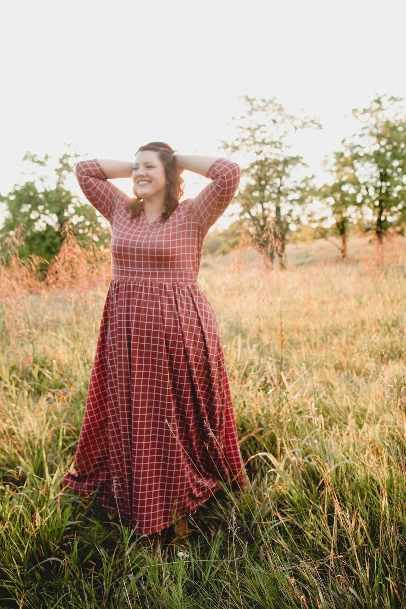 Woman wearing modest copper windowpane plaid maxi dress with 3/4 sleeves and v-neckline, standing gracefully in outdoor field setting with trees in background, demonstrating the dress's comfortable fit and nursing-friendly design