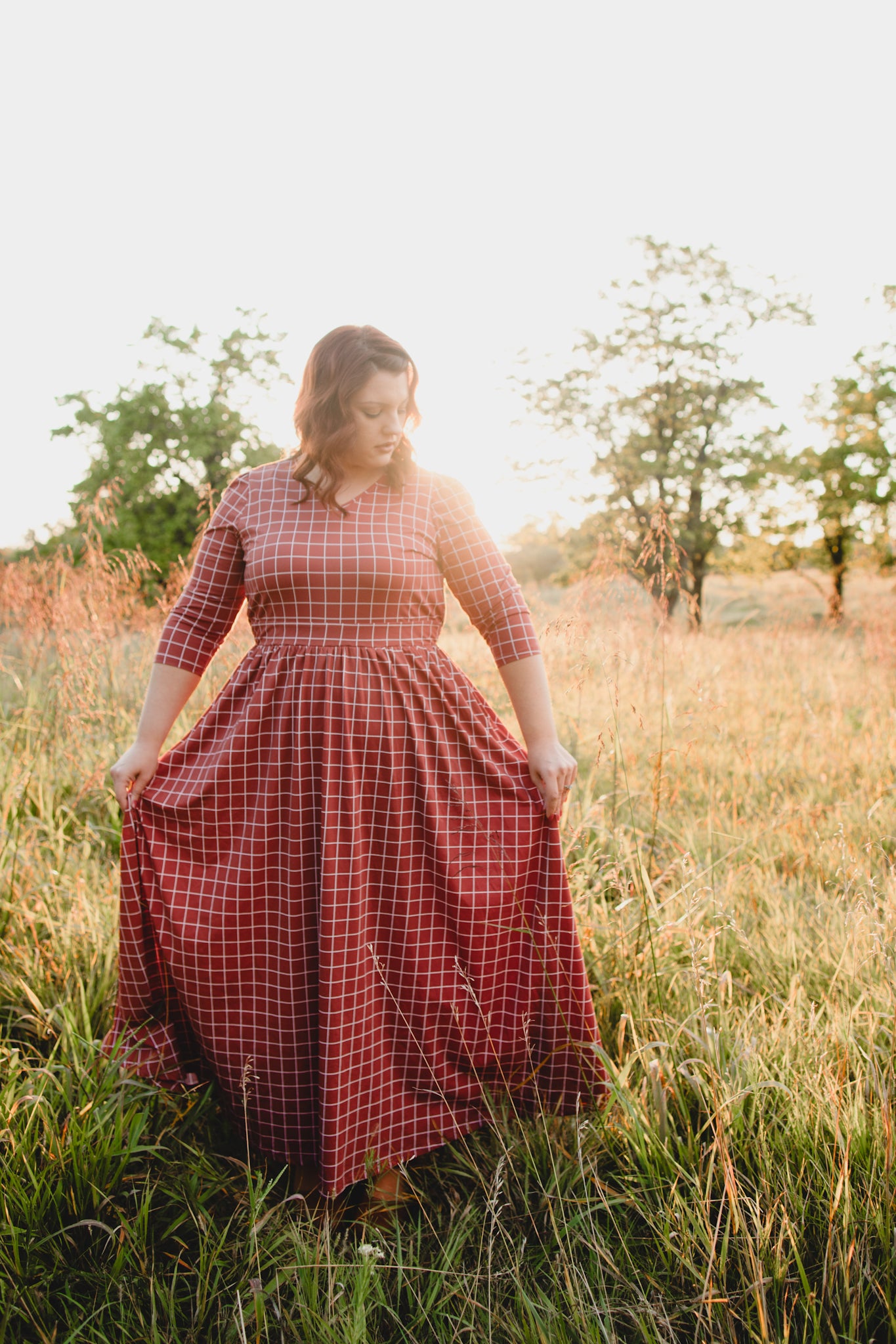 Woman wearing modest copper windowpane plaid maxi dress with 3/4 sleeves and v-neckline, standing gracefully in outdoor field setting with trees in background, demonstrating the dress's comfortable fit and nursing-friendly design