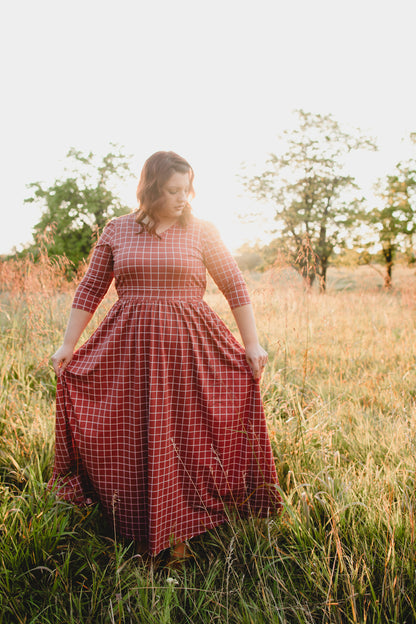 Woman wearing modest copper windowpane plaid maxi dress with 3/4 sleeves and v-neckline, standing gracefully in outdoor field setting with trees in background, demonstrating the dress's comfortable fit and nursing-friendly design