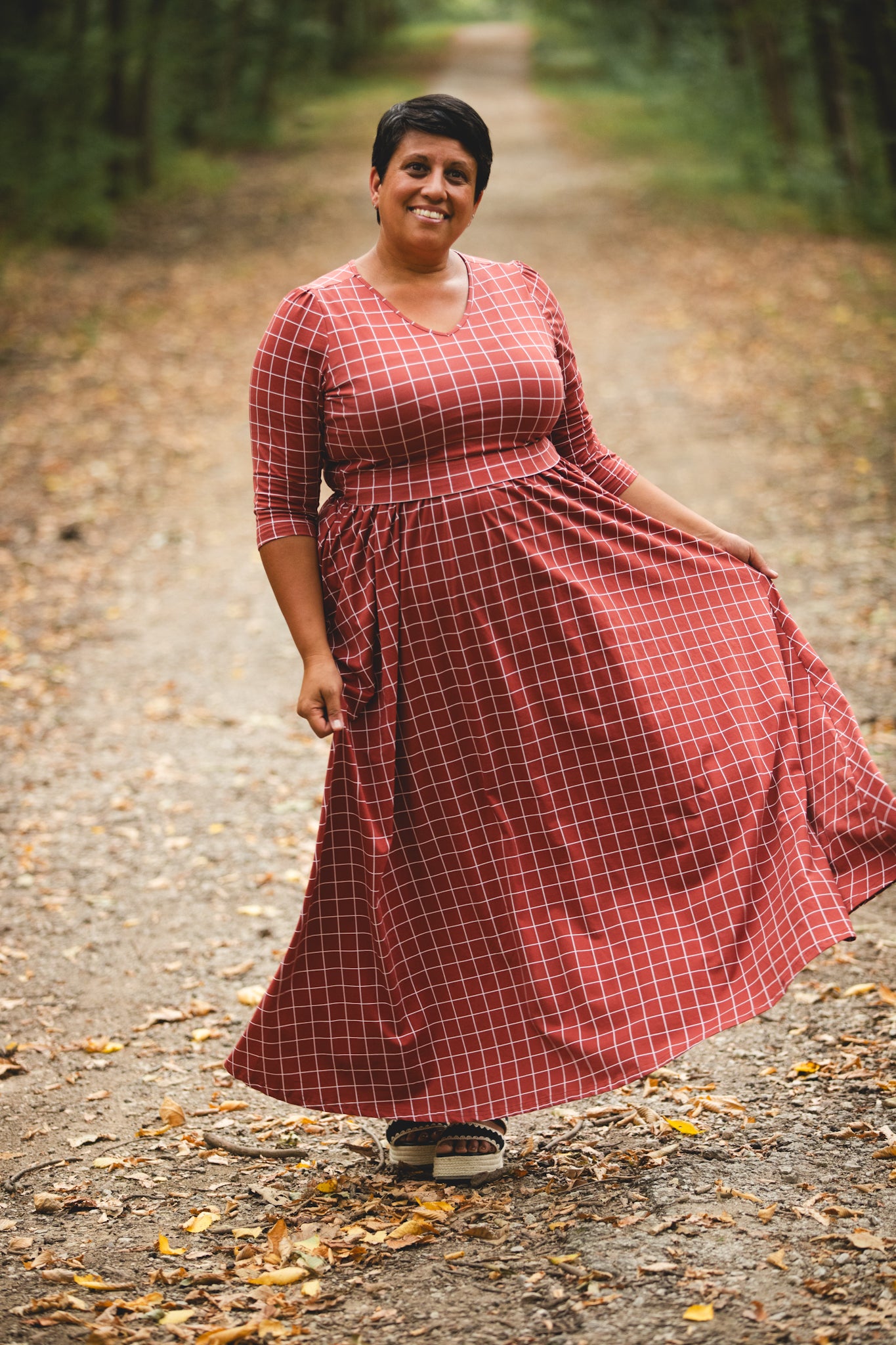 Woman wearing modest copper windowpane plaid maxi dress with 3/4 sleeves and v-neckline, standing confidently on forest pathway surrounded by autumn trees, showcasing the nursing-friendly design and comfortable fit perfect for nature walks and outdoor adventures