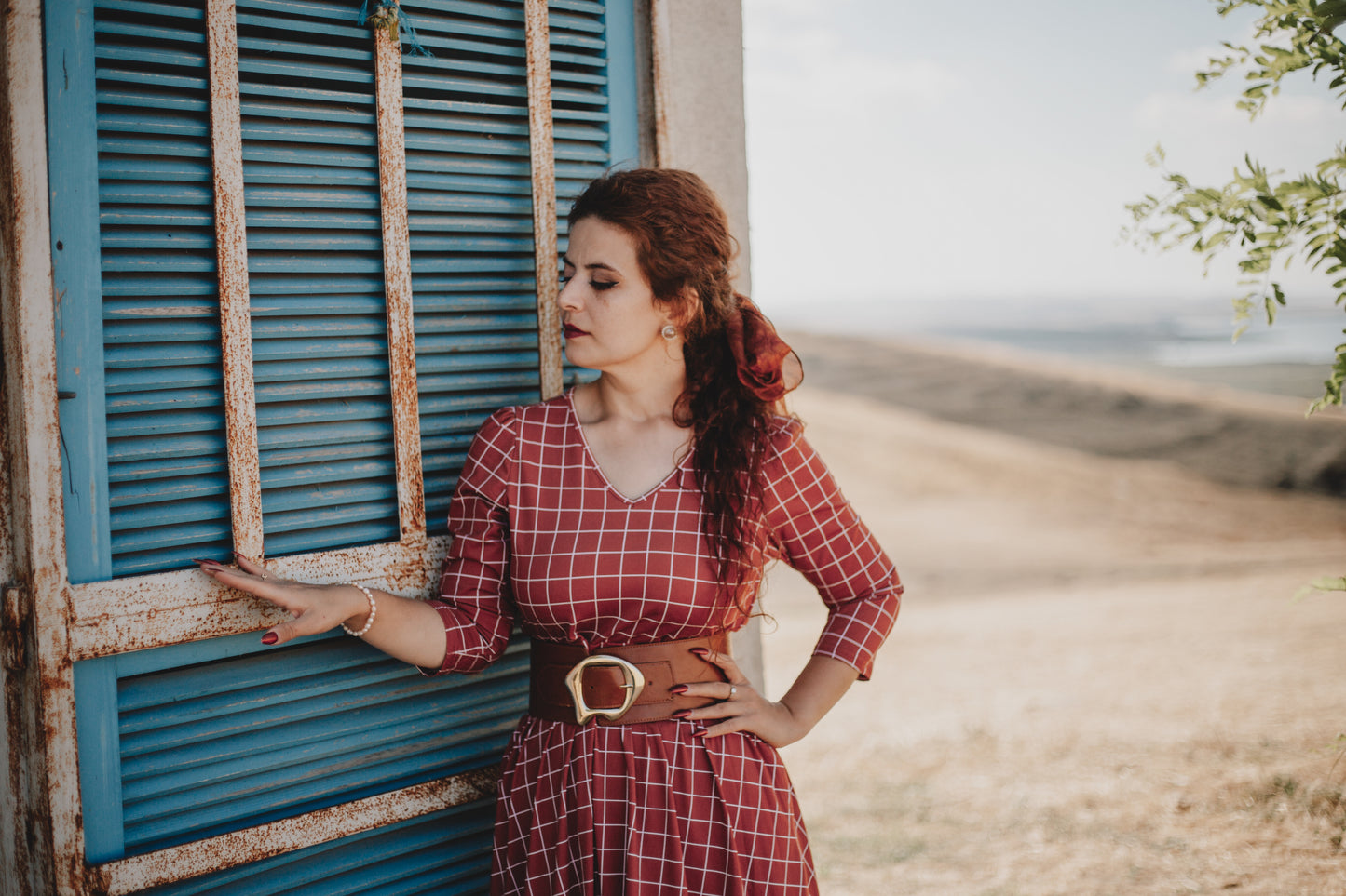 Woman wearing modest copper windowpane plaid maxi dress with 3/4 sleeves and v-neckline, standing elegantly by rustic blue shuttered door, demonstrating the nursing-friendly design and versatile cotton spandex style perfect for casual and formal occasions
