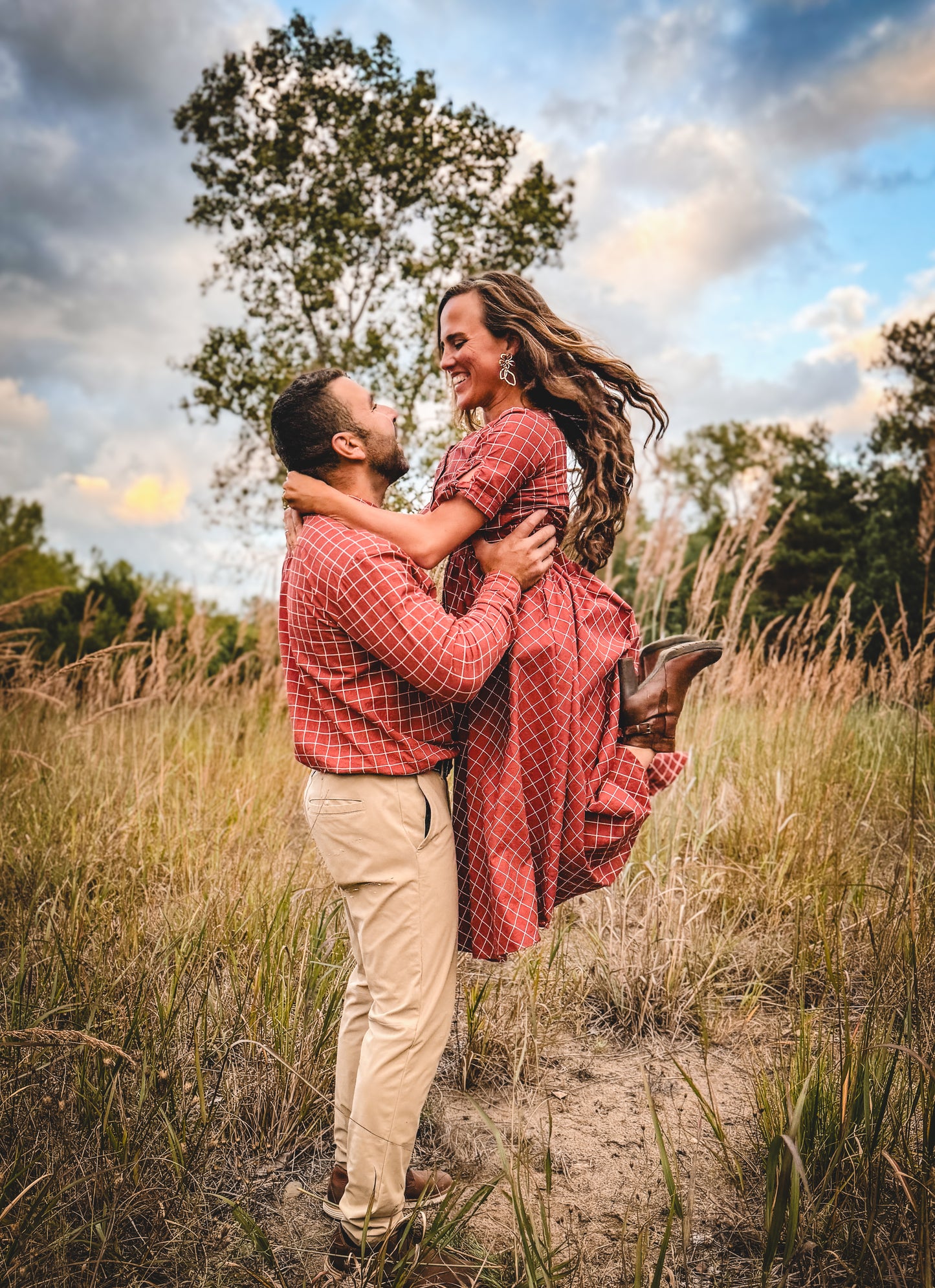 Couple in red plaid shirts embracing in a field with a blue sky and trees.