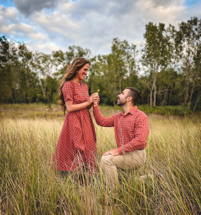 Man proposing to a woman in a red dress in a grassy field with trees in the background