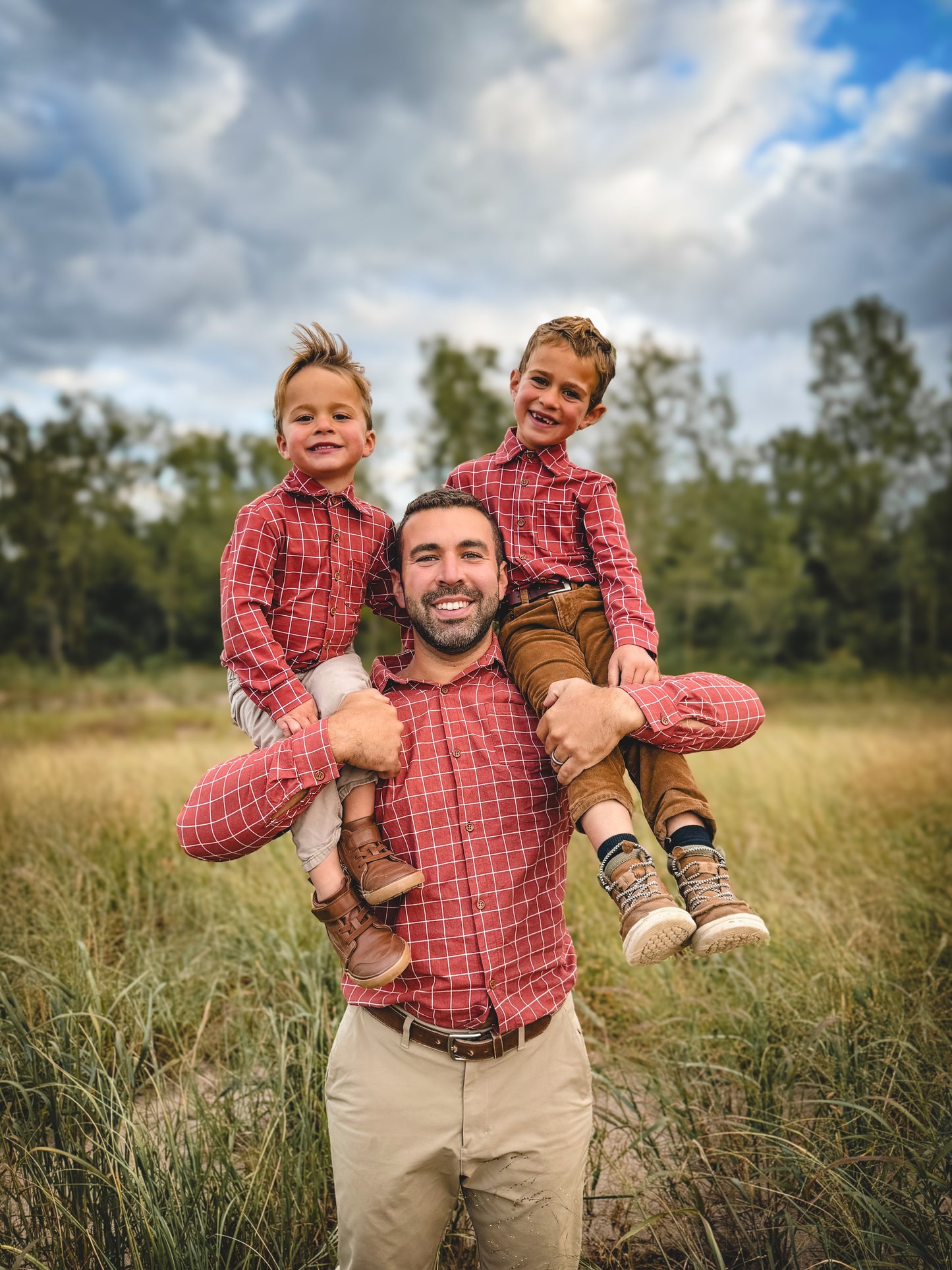 Man with two children on his shoulders in a field with trees and blue sky.