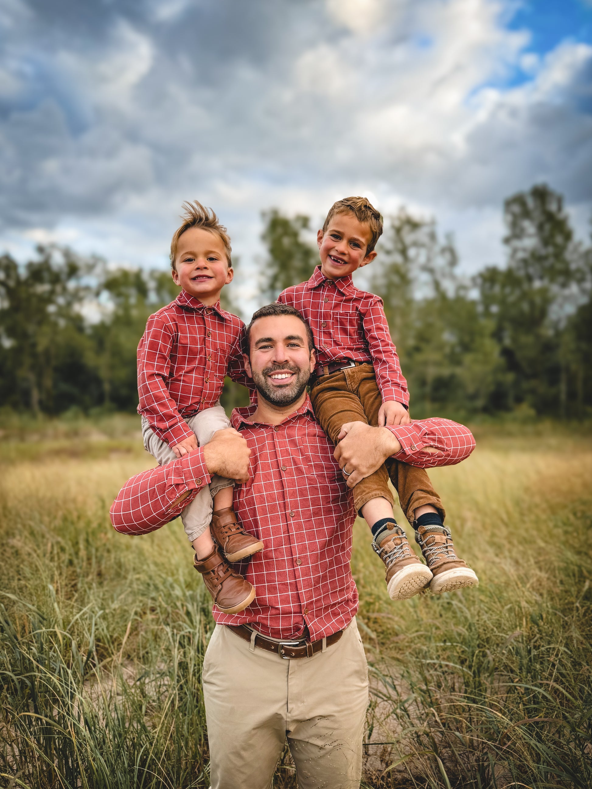 Man with two children on his shoulders in a field with trees and blue sky.