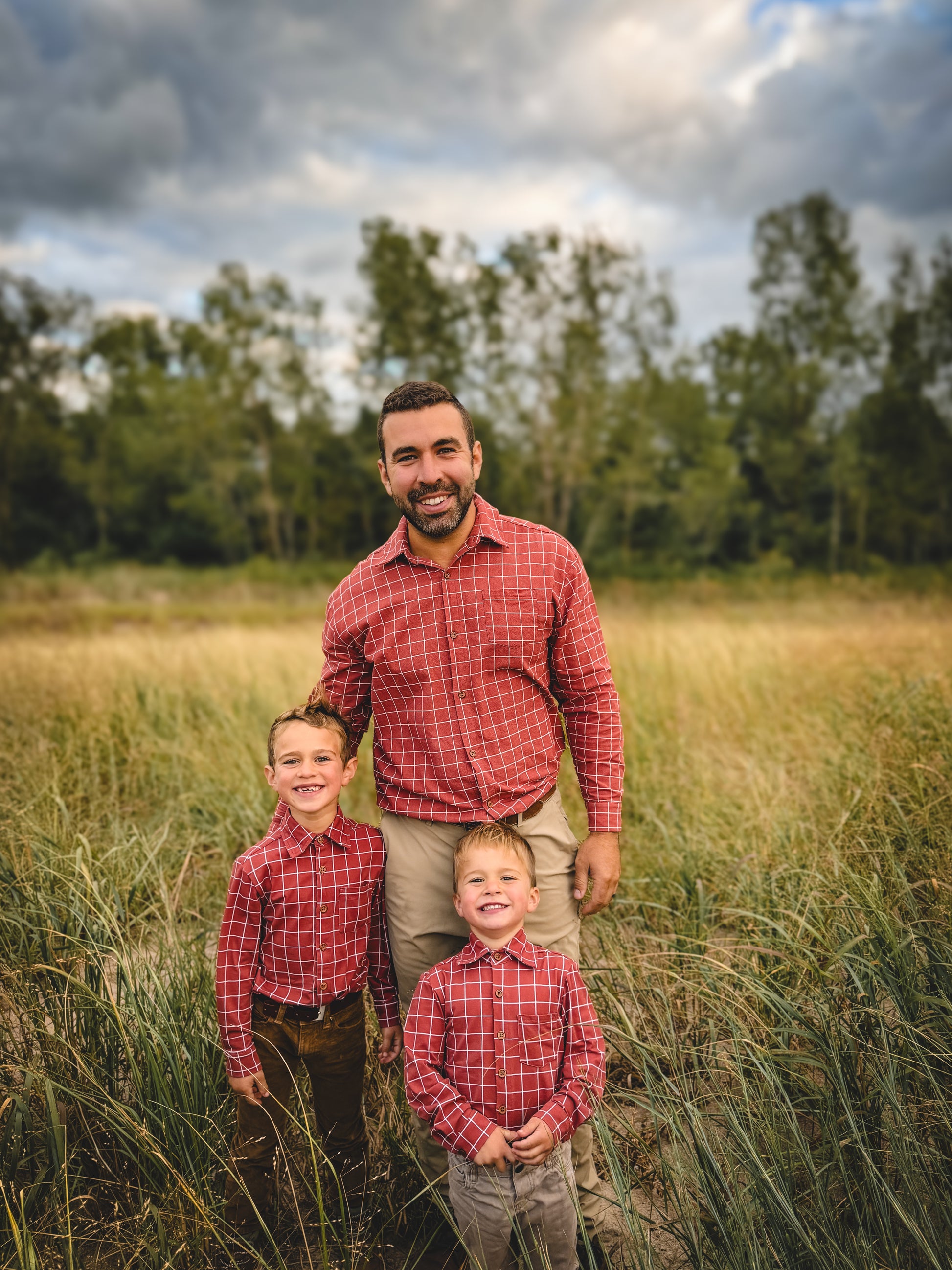 Man and two children in matching red plaid shirts standing in a grassy field with trees in the background.