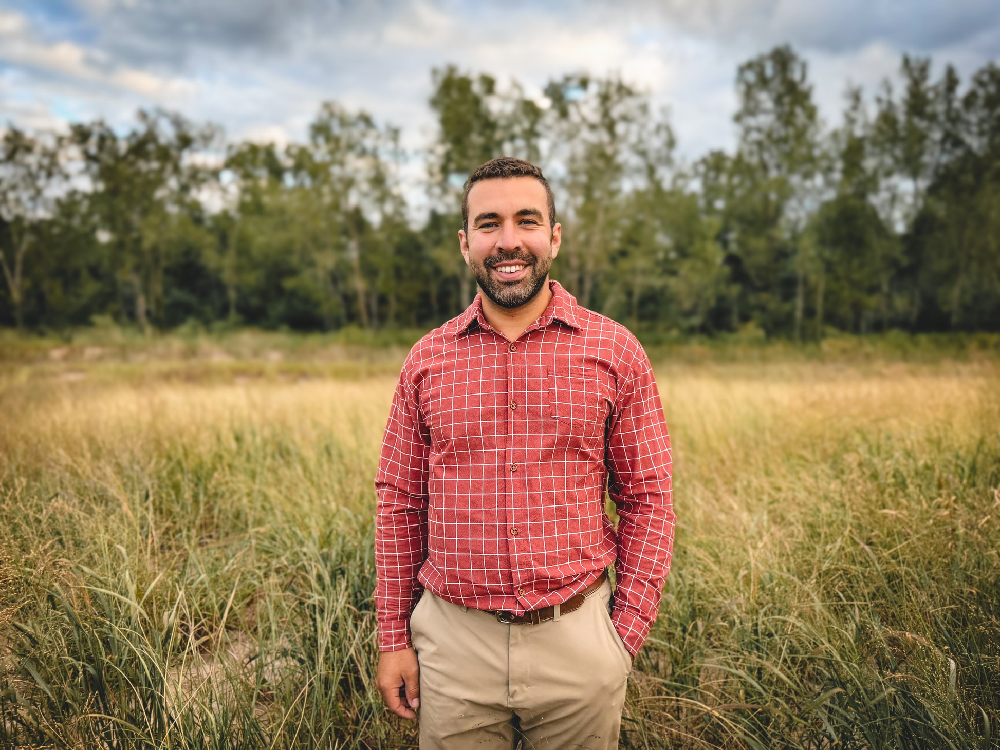 Man standing in a field wearing a red plaid shirt and beige pants.