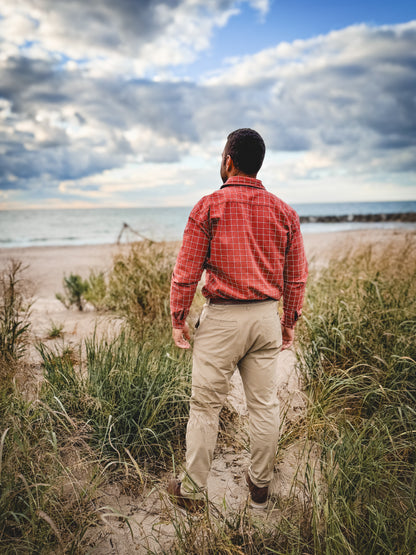 Man in a red plaid shirt and beige pants walking along a beach path with a cloudy sky.