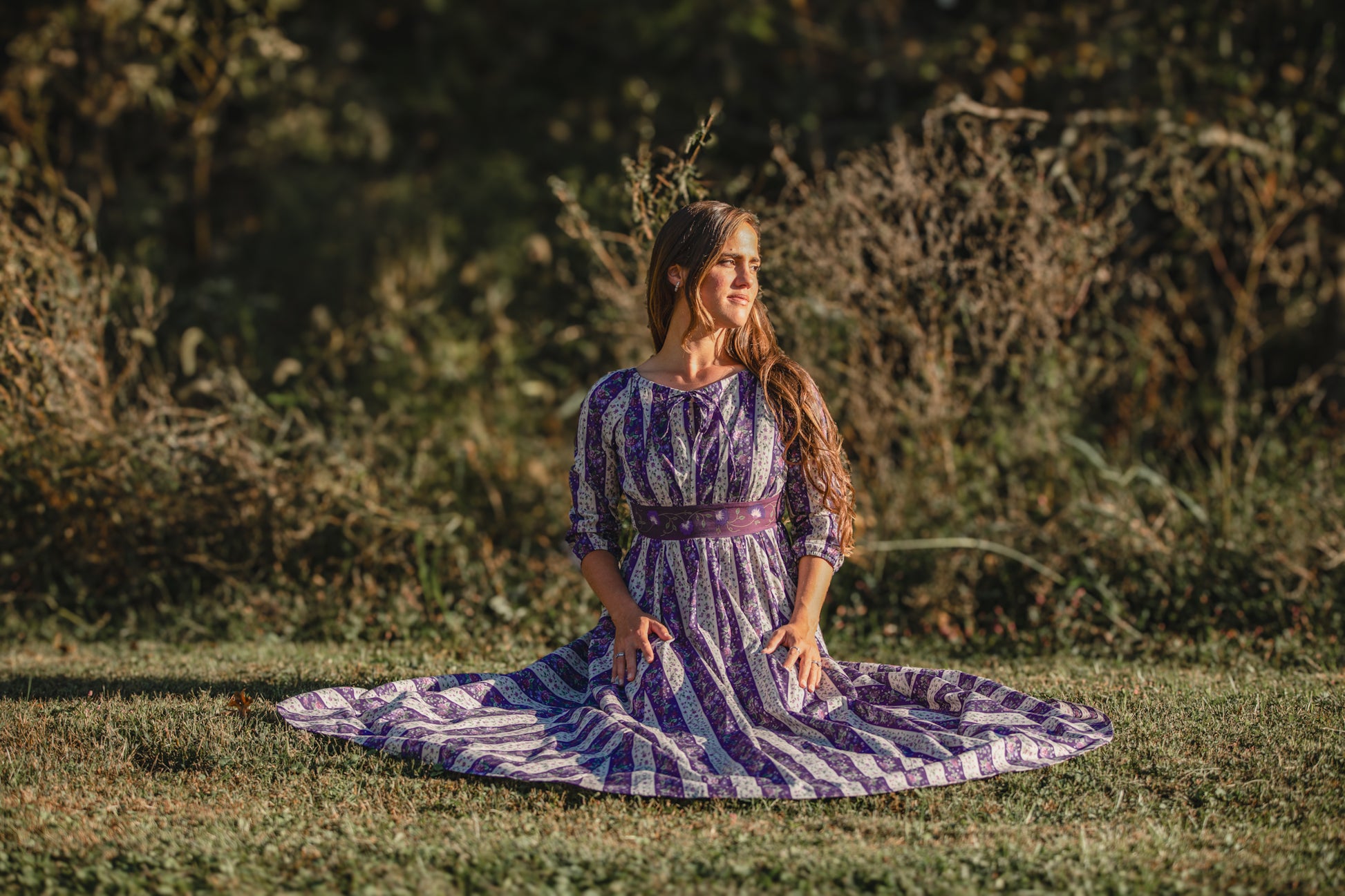 Woman in a purple dress standing in a grassy field with trees in the background