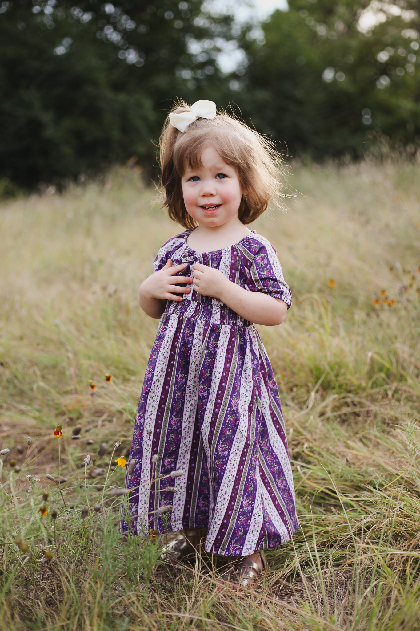 Young girl in a purple dress standing in a grassy field with trees in the background
