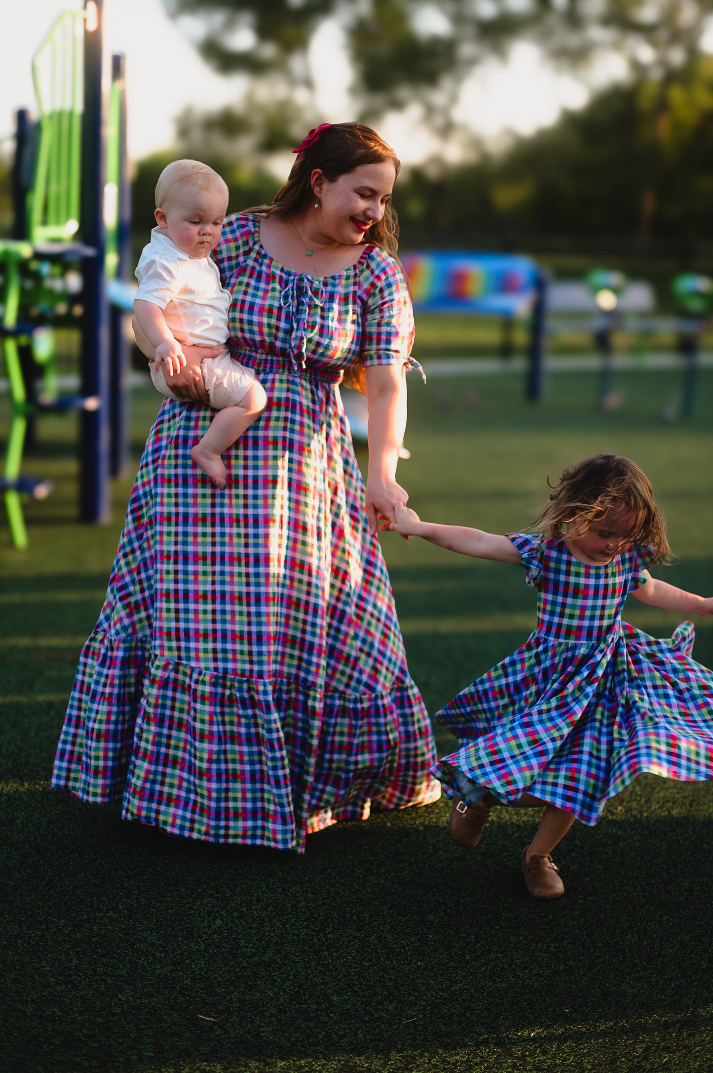Mother and daughter in plaid modest dresses holding a baby and walking with a child in a similar dress on grass.