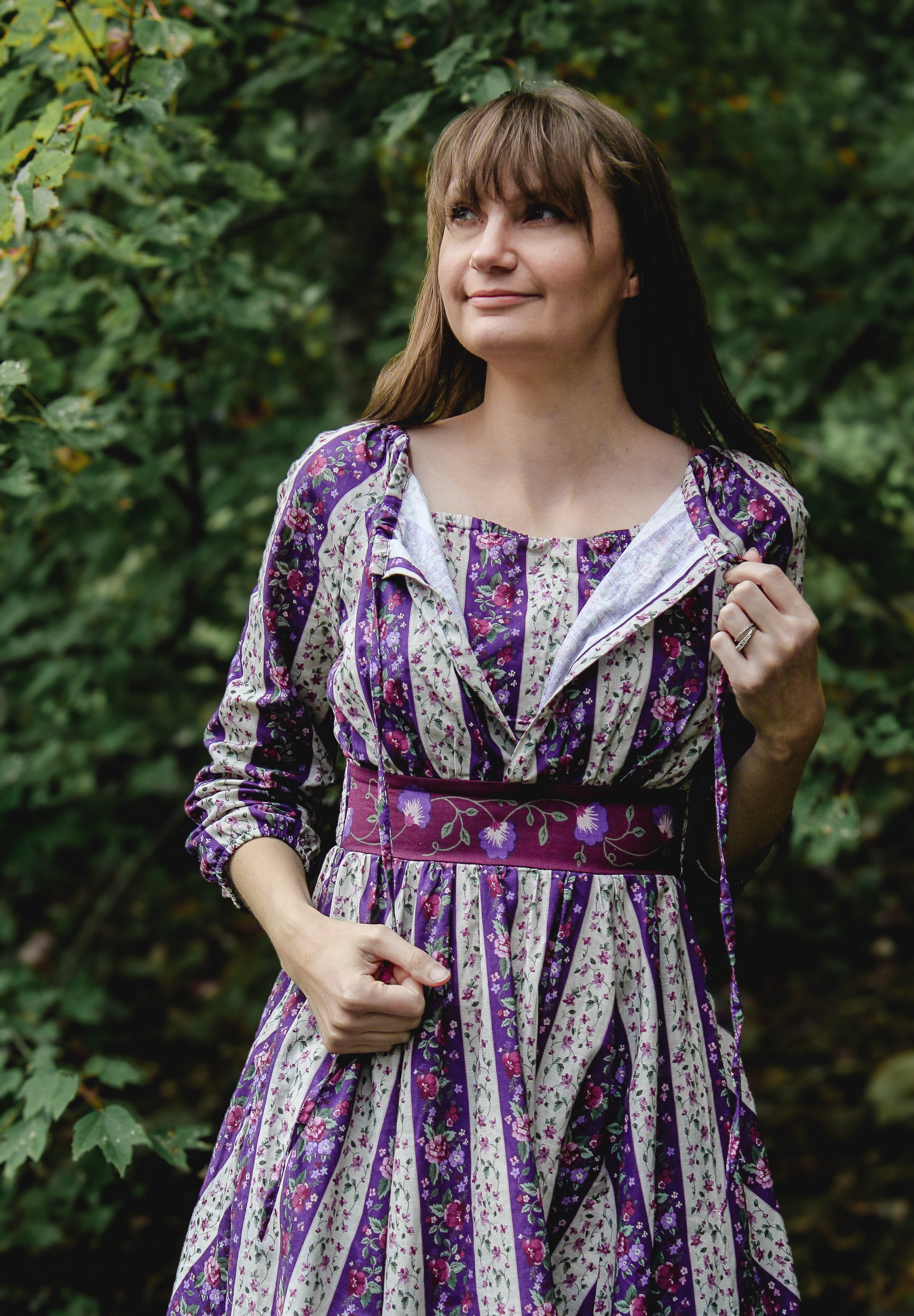 Woman wearing a modest nursing purple floral dress standing in front of green foliage