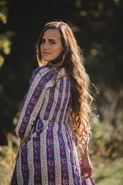 Woman in a striped modest nursing dress standing outdoors with trees in the background