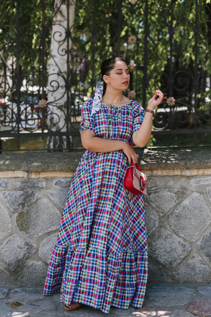 Woman in a colorful plaid modest nursing dress standing outdoors with a stone wall and trees in the background.