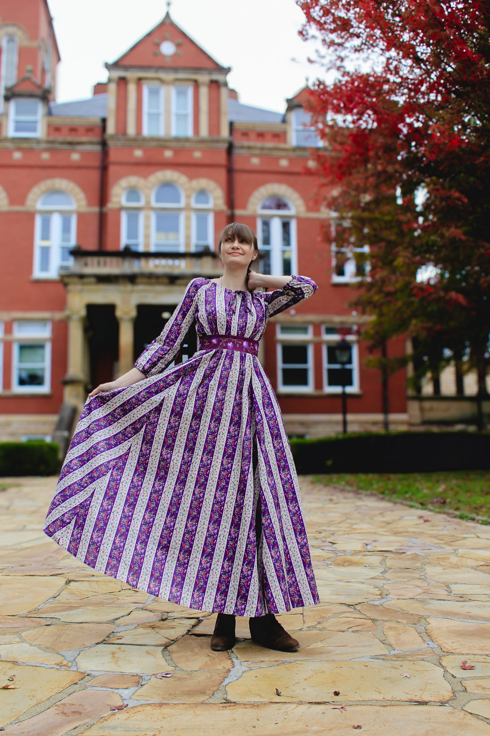 Woman in a modest nursing purple and white striped dress standing in front of a red brick building with trees in the background.